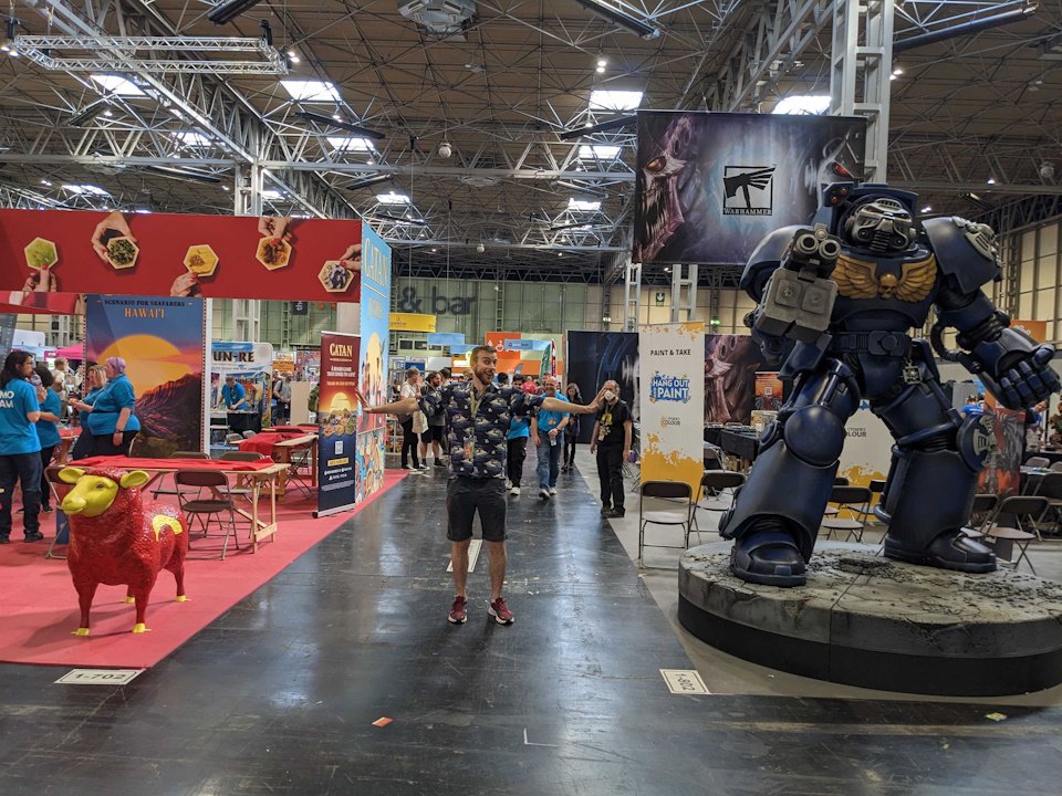 UK game expo non award awards rivalry catan warhammer - a man stands with arms outstretched between a catan sheep figure and large terminator model