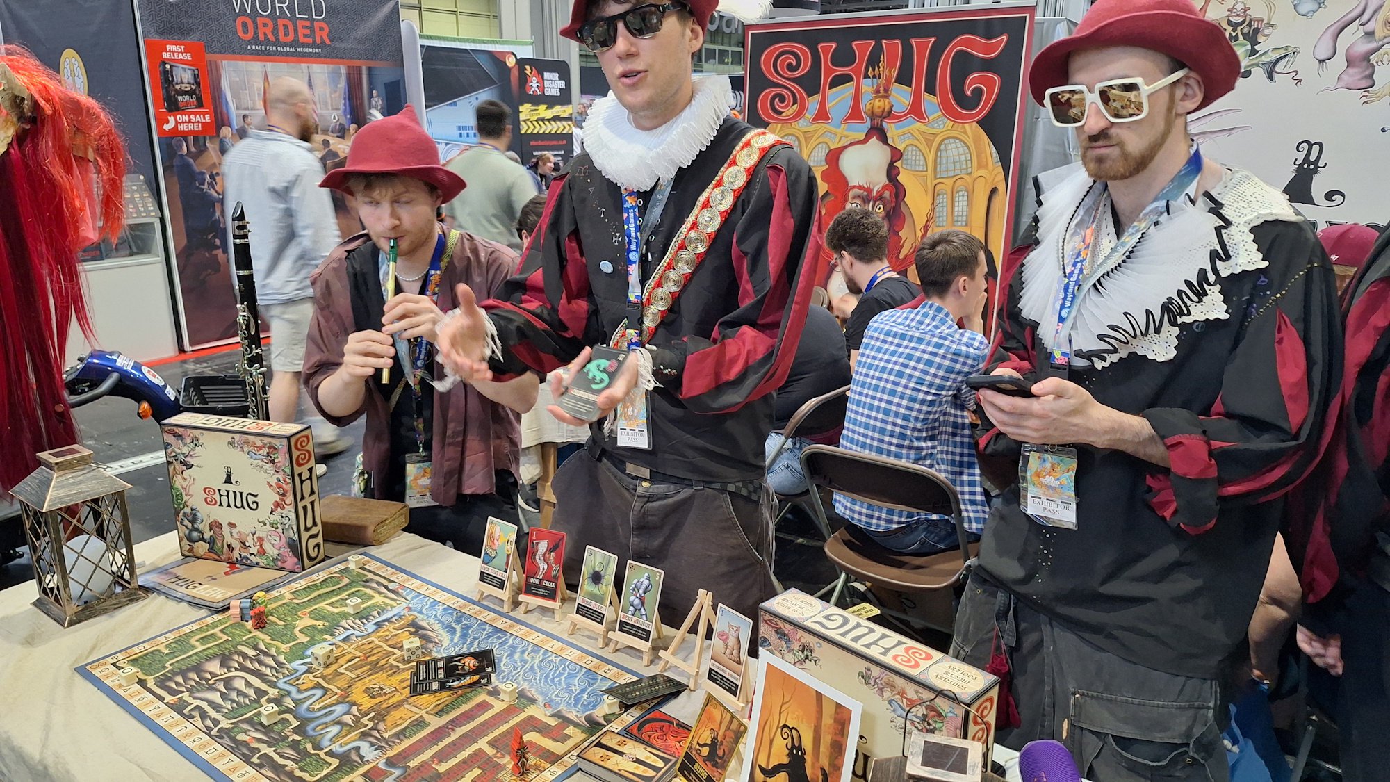 Three men dressed as medieval bard wearing sunglasses in front of the baord game SHUG. One plays a flute while the other explains